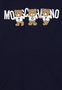 Námořnická modrá trička s třemi kreslenými medvědy a textem "Moschino Toy" v tučných bílých písmenkách nad nimi. Medvědi mají na sobě bílé košile.