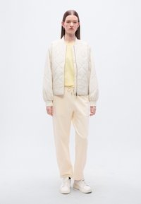 Κρεμ μπουφάν bomber με καπιτονέ σχέδιο και ελαστικές μανσέτες, συνδυασμένο με κίτρινο τοπ και χλωμό μπεζ παντελόνι φόρμας. Λευκά αθλητικά παπούτσια ολοκληρώνουν την εμφάνιση.