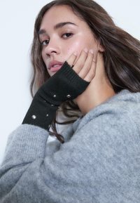 Guanti neri senza dita realizzati in tessuto a coste, con borchie argentate al polso, abbinati a un maglione grigio chiaro.