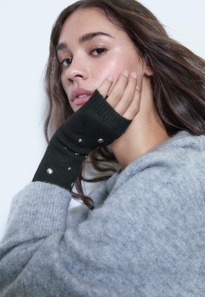 Schwarze fingerlose Handschuhe aus geripptem Strickstoff, mit silbernen Nieten am Handgelenk, kombiniert mit einem hellgrauen Pullover.