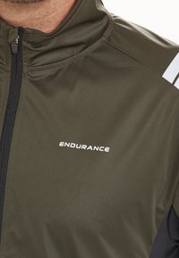 Giacca sportiva verde oliva realizzata in tessuto liscio, con chiusura a zip, accenti bianchi riflettenti sulle spalle e logo "ENDURANCE" in bianco.