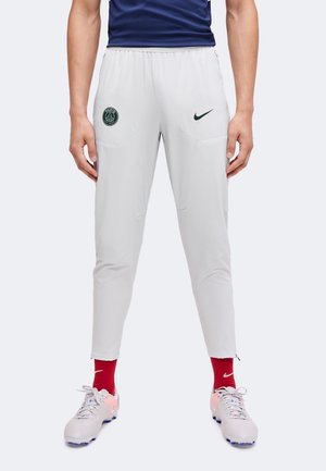 Άτομο που φοράει λευκό αθλητικό παντελόνι Nike με το λογότυπο της Paris Saint-Germain, κόκκινες κάλτσες Nike και ανοιχτό γκρι παπούτσια ποδοσφαίρου, στέκεται με ανοιχτά πόδια.