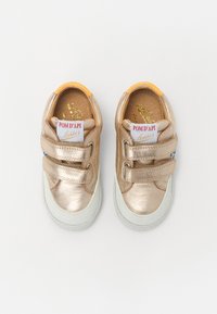 Baskets métalliques dorées avec deux sangles Velcro, semelle en caoutchouc blanche et accents orange au niveau du talon. Affiche l'étiquette de la marque à l'intérieur de la chaussure.