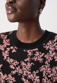 Črna pulover z rožnatim cvetličnim vzorcem, okrogla rebrasta ovratnica in gladka tekstura. Pogleduj bližnji pogled na izrez in podrobnje ramena.