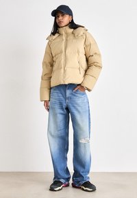 Giacca trapuntata beige con colletto alto, chiusura a zip e tasche laterali. Indossata con jeans a gamba larga in blu sbiadito e sneakers nere con dettagli rosa.