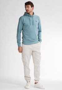 Sudadera azul claro con un bolsillo delantero y cordones, combinada con pantalones cargo beige que tienen múltiples bolsillos, y zapatillas blancas.