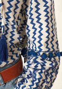 Bílá látková košile se modrým cik-cak vzorem, doplněná modrými bambulkami podél manžet a třásněmi po straně. Viditelný hnědý kožený pás.