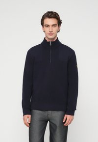 Pull-over bleu marine avec col montant, fermeture éclair au quart, texture côtelée et panneaux d'épaule contrastants. Porté avec un pantalon gris.