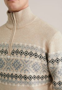 Beżowy sweter dzianinowy z wysokim kołnierzem, z geometrycznym wzorem w odcieniach szarości i czerni na klatce piersiowej i ramionach. Zamek błyskawiczny.