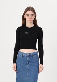 Schwarzes, geripptes Cropped-Top mit langen Ärmeln. Mit weißem Logo-Text auf der Brust. Kombiniert mit einem blauen Denim-Bleistiftrock.