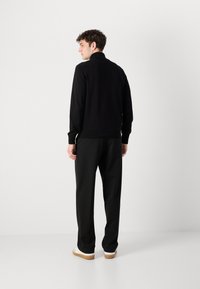 Svart stickad tröja med hög krage, kombinerad med svarta byxor och vita sneakers. Modellen sedd från ryggen, vilket visar en avslappnad look.