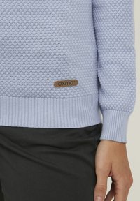 Svetlo modra teksturirana jopica z rebrastimi robovi in manšetami, z rjavim odtisom OXMO na spodnji strani. V kombinaciji z črnimi hlačami.