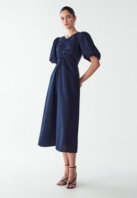 Mørkeblå kjole med puffærmer, indsnævret talje og en løs, flydende nederdel. Fremstillet af struktureret stof med snøredetalje.