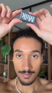 Man with mustache and goatee holds black rectangle labeled "Milk" above his head in a room with wooden shelves and green plants.