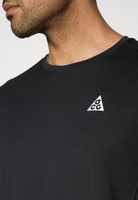 Μαύρο μπλουζάκι κατασκευασμένο από λείο ύφασμα, με λευκό τριγωνικό λογότυπο στο στήθος. Στρογγυλή λαιμόκοψη και κοντά μανίκια.