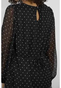 Blouse noire à pois avec manches longues transparentes, taille nouée et ouverture en trou de serrure dans le dos, ornée de pois blancs circulaires sur toute la surface.
