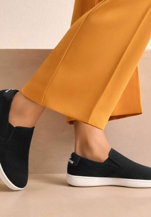 Black slip-on shoes with a textured upper, white rubber sole, and a small logo on the heel. Paired with a yellow wide-leg pant.