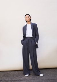 Grauer maßgeschneiderter Blazer mit weit geschnittenen Hosen, kombiniert mit einem weißen T-Shirt und hellen Schuhen. Glattes Material und strukturierte Form.