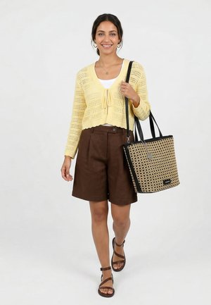 Hymyilevä nainen kävelee, hänellä on keltainen neuleneuletakki, valkoinen toppi, ruskeat shortsit, ruskeat sandaalit ja iso kudottu kangaskassi mustilla hihnoilla.