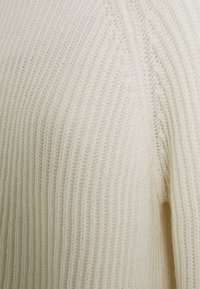 Pull en maille de couleur blanc cassé, avec une texture côtelée, un col rond et une coupe légèrement surdimensionnée. Tissu doux avec des détails de couture visibles.