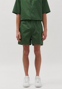 Set di camicia a maniche corte e pantaloni corti abbinati di colore verde, realizzato in tessuto liscio. La camicia ha una tasca; i pantaloni corti presentano una cintura elastica. Sneakers bianche.