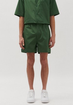 Set di camicia a maniche corte e pantaloni corti abbinati di colore verde, realizzato in tessuto liscio. La camicia ha una tasca; i pantaloni corti presentano una cintura elastica. Sneakers bianche.