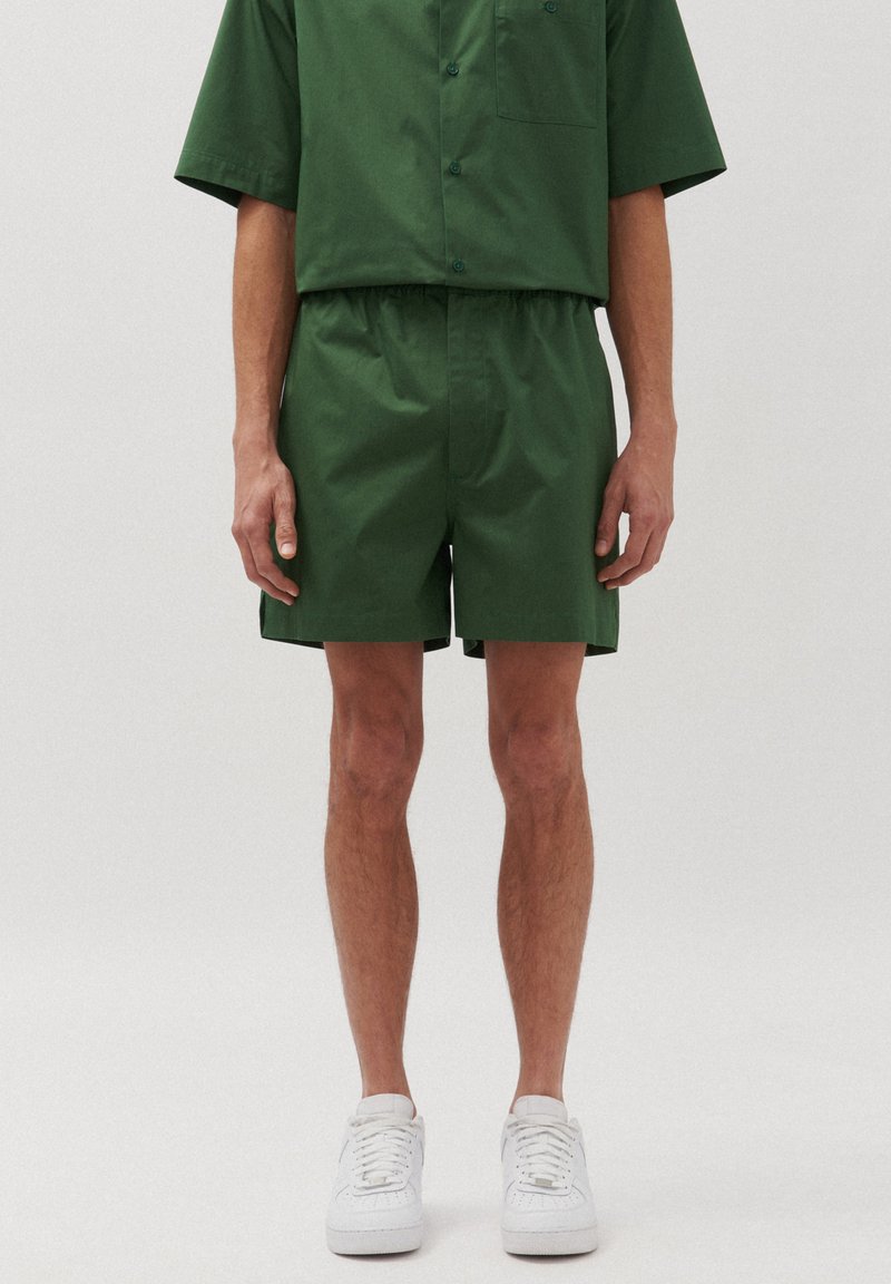 Set di camicia a maniche corte e pantaloni corti abbinati di colore verde, realizzato in tessuto liscio. La camicia ha una tasca; i pantaloni corti presentano una cintura elastica. Sneakers bianche.