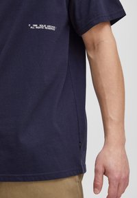 Marineblå bomulds T-shirt med korte ærmer, rund hals og et lille hvidt labeldetalje nær kanten med teksten "C 1999 SOLID ARCHIVE."