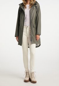 Olivově zelený pláštěnka s kapucí, s předními kapsami a stříbrnými detaily, nošená přes pruhovanou košili a krémové kalhoty, doplněná béžovými botami.