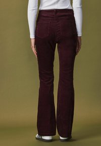 Bordeaux velour flared bukser med baglommer, båret med en hvid top og sort fodtøj, sat mod en dæmpet grøn baggrund.