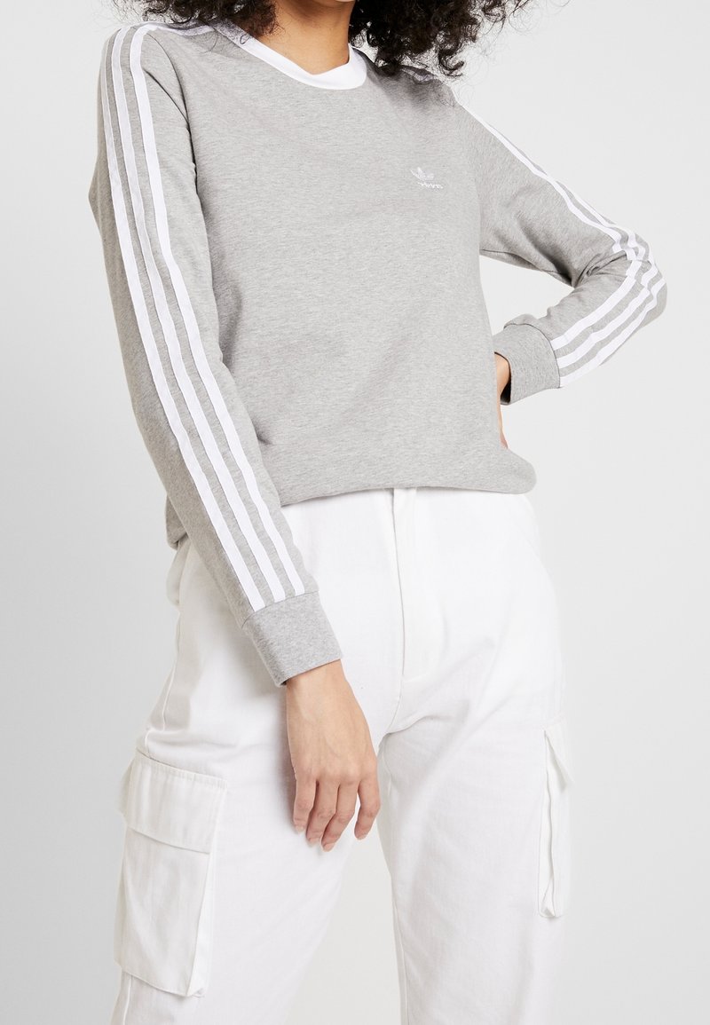 Grå sweatshirt med vit tre-stripesdesign på ärmarna och en vit krage. Tillverkad av mjukt material, med en diskret logotyp på bröstet.