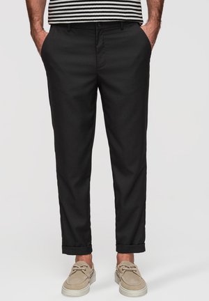Homme portant un pantalon noir à revers, des chaussures décontractées beiges et une chemise rayée, les mains dans les poches, sur un fond uni.