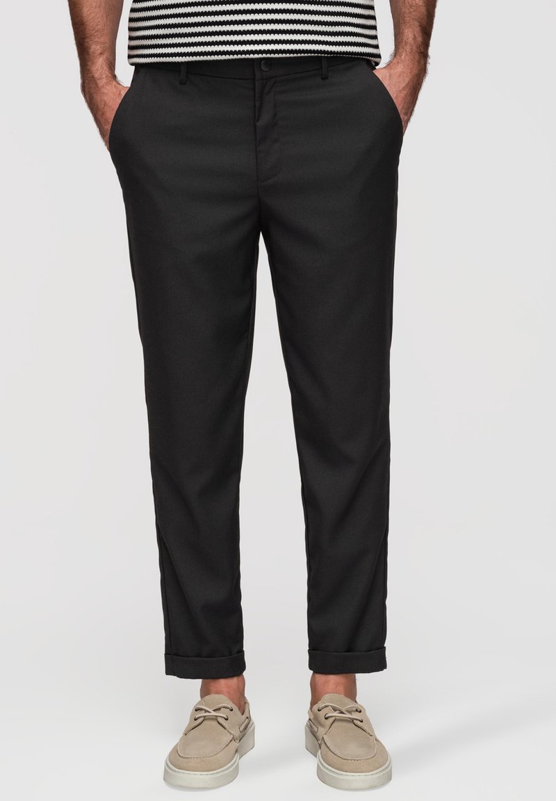 Homme portant un pantalon noir à revers, des chaussures décontractées beiges et une chemise rayée, les mains dans les poches, sur un fond uni.
