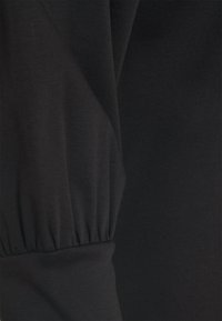 Černá tkanina se shromážděným elastickým detailem vytvářejícím jemné záhyby a texturu, naznačující rukáv nebo manžetu oděvu.