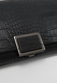 Pochette noire en cuir synthétique façon crocodile avec une boucle rectangulaire argentée brillante sur une surface blanche.