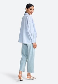 Gestreept blauw shirt met kraag, gecombineerd met lichte denim broeken en witte slingback hakken. Model poseert van achteren en toont de details van de outfit.