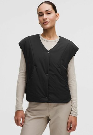 Naine, kes kannab musta varrukateta polsterdatud vesti beeži pikavarrukalise pluusi ja beežide pükstega, seisab ühevärvilise heleda tausta ees.