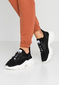 Baskets en mesh noires avec une semelle blanche, dotées d'accents noirs et de lacets élastiques. La chaussure a un bout arrondi et une surface texturée.