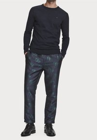 Mężczyzna w czarnej longsleeve'owej bluzie, granatowych spodniach w zielony wzór palm, czarnych eleganckich butach, stojący z rękami w kieszeniach, na białym tle.