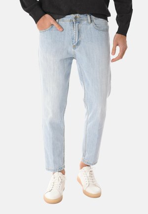 Persona che indossa jeans azzurri con le mani parzialmente nelle tasche e sneakers bianche con suole in gomma, su uno sfondo neutro.