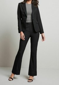 Schwarze maßgeschneiderte Sakko und ausgestellte Hose, kombiniert mit einem grauen gerippten Oberteil. Die Schuhe sind schwarze, geschnürte High Heels. Glattes Material, strukturiertes Design.