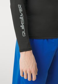 Svart langermet rash guard med "Quiksilver"-logoen i lys grå på venstre arm, kombinert med blå badetights. Glatt tekstur.