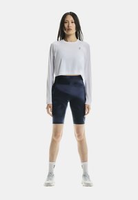 Donna in piedi di fronte, indossa un crop top bianco a maniche lunghe, pantaloncini da bici blu navy, calzini bianchi e scarpe da ginnastica bianche su uno sfondo bianco.