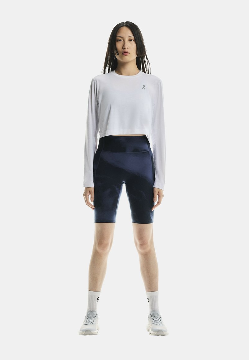 Donna in piedi di fronte, indossa un crop top bianco a maniche lunghe, pantaloncini da bici blu navy, calzini bianchi e scarpe da ginnastica bianche su uno sfondo bianco.