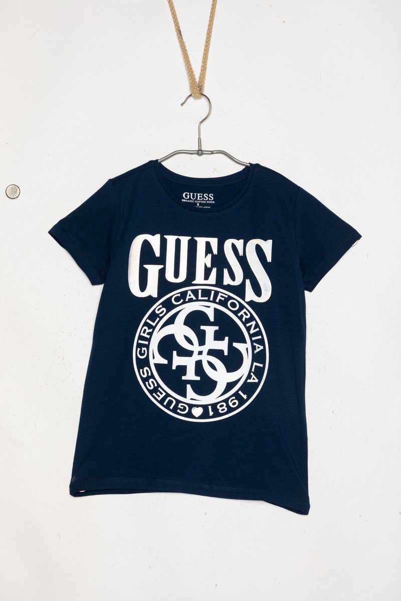 Námorně modré bavlněné tričko s velkými bílými nápisy "GUESS" a kulatým logem s textem. Krátké rukávy, kulatý výstřih a měkká textura.