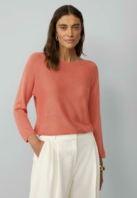 Koralewe sweter robiony na szydełku z okrągłym dekoltem i długimi rękawami, zestawiony z białymi plisowanymi spodniami. Materiał wydaje się lekki i lekko fakturowany.