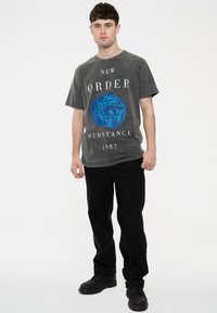 Gray cotton t-shirt with front graphic design, featuring "NEW ORDER SUBSTANCE 1987" text and a blue abstract shape, paired with black pants.