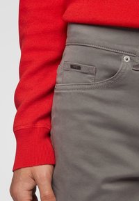 Pantalones de algodón grises con costuras tonales, que presentan un bolsillo frontal con una etiqueta de marca y un cierre de botón. Destacados por la manga de un suéter rojo.