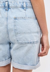 Κοντινό πλάνο ενός ατόμου που φοράει ανοιχτό μπλε τζιν σορτς με μεγάλη πίσω τσέπη και γυρισμένες άκρες, με το χέρι να ακουμπά στο πλάι.