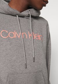 Felpa grigia con logo "Calvin Klein" rosa, cappuccio con cordino, estremità metalliche sui cordini e tasca anteriore. Tessuto morbido.